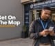 A Botswana business owner standing outside a commercial building holding a smartphone showing a map location marker.